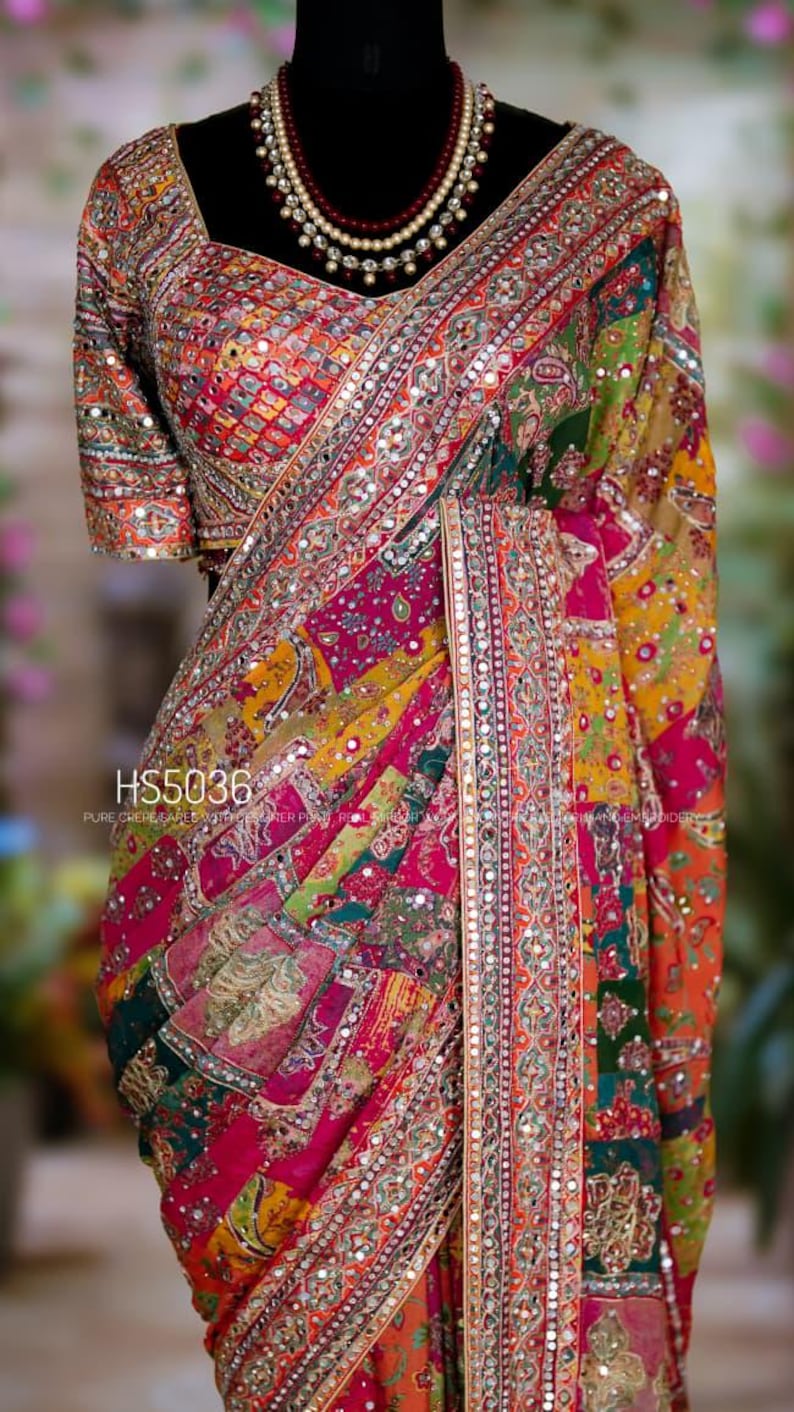 May include: A vibrant, multi-colored saree with intricate embroidery and mirror work. The design features a patchwork pattern in shades of pink, orange, green, and yellow. The text "HS5036" is visible on the fabric. A beaded necklace is also visible.