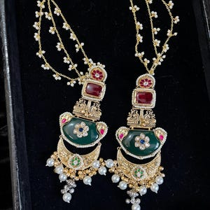 May include: A pair of ornate gold earrings with a dark green, emerald-shaped center stone. The earrings feature red and blue accents, small white pearls, and a chain of small white flowers. The earrings are displayed on a black surface.