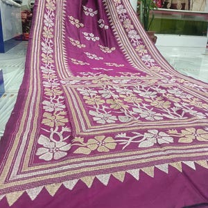 May include: A vibrant magenta textile with intricate floral embroidery. The design features cream and gold-colored flowers and geometric patterns along the borders. The fabric is draped, showcasing the detailed craftsmanship and texture.