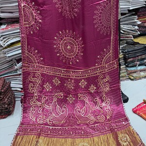 Puede incluir: Un textil magenta con diseños circulares y de pavo real dorados. La tela presenta patrones intrincados y un borde dorado. El textil es probablemente una bufanda o chal, que muestra el arte tradicional y los colores vibrantes.
