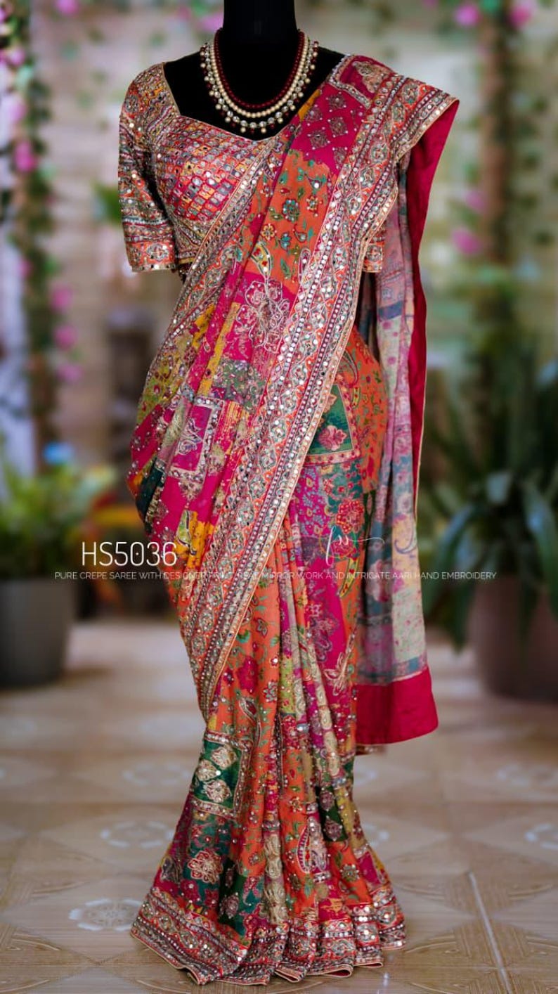 May include: A vibrant, patterned saree in shades of orange, pink, and green. The fabric is adorned with intricate embroidery and embellishments. The text "HS5036" is visible on the saree. The saree is draped on a mannequin.
