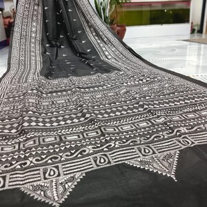 Könnte beinhalten: Schwarzer Stoff mit aufwendigen weißen Stickmustern. Das Design zeigt geometrische Formen, florale Motive und detaillierte Bordüren. Der Stoff ist drapiert und zeigt die detaillierte Handwerkskunst und Textur des Materials.