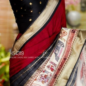 May include: A black and red saree with gold accents and a detailed border. The saree features a decorative border with a cream-colored panel with illustrations of people and vehicles. The bodice is black with gold embellishments.