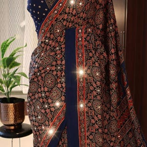 May include: A navy blue saree with a detailed paisley pattern in red and black. The saree features a decorative border and is embellished with small, reflective accents. The design includes a matching navy blue blouse.