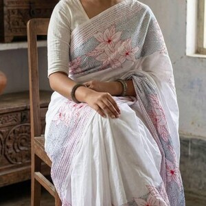 May include: A white saree with a floral pattern in red and gray, worn with a matching white blouse. The saree features intricate embroidery and is draped over a person seated on a wooden chair.