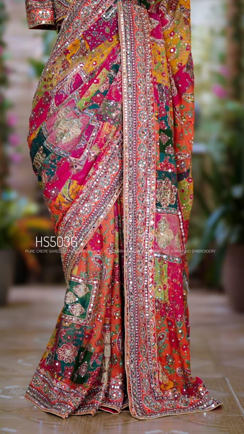 May include: A multi-colored crepe saree featuring a patchwork design in shades of pink, orange, yellow, and green with intricate Aari hand embroidery and real mirror work. The fabric displays the text HS5036 PURE CREPE SAREE.