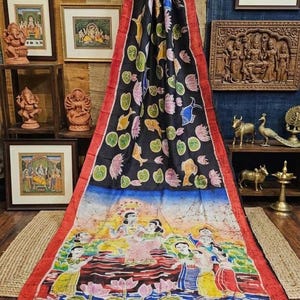 May include: A black and red sari with a colorful painted design. The top portion features floral and fish motifs, while the bottom depicts a scene with figures in a water setting. The sari is displayed on a wooden surface.