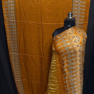 May include: Mustard-colored fabric with a decorative border and intricate patterns. The fabric is draped over a mannequin, showcasing the design. The border features a repeating pattern of teardrop shapes and circles.