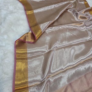 Pode incluir: Um tecido brilhante, castanho claro, com uma borda dourada e acabamento rosa. O tecido tem um padrão sutil e texturizado e um acabamento brilhante. A borda dourada adiciona um toque de elegância. Adequado para lenços, xales ou outras peças de vestuário.