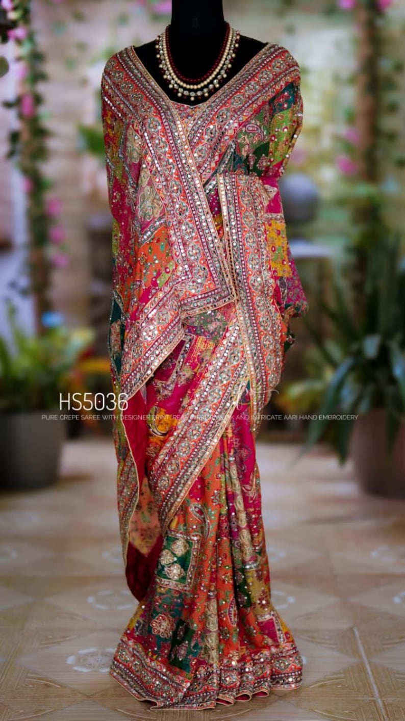 May include: A multi-colored crepe saree featuring intricate Aari hand embroidery, mirror work, and a wide decorative border. The fabric displays a vibrant pattern of pink, orange, and green. Text on the image reads HS5036 PURE CREPE SAREE.