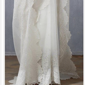 May include: A white wedding dress with intricate lace detailing. The dress features a flowing skirt with a scalloped edge and delicate floral patterns. The fabric appears sheer and lightweight, creating an elegant and romantic look.