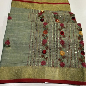 May include: A folded textile with a sage green base, featuring embroidered floral patterns in red, orange, and yellow. The fabric has a gold border with a red trim. The design includes vertical stitched lines and scattered sequins.