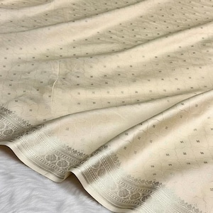 Può includere: Tessuto color avorio con una sottile lucentezza e un motivo ripetuto di piccoli motivi quadrati. Il tessuto ha un bordo decorativo con intricati disegni floreali e geometrici in una tonalità grigio argento. Il tessuto è drappeggiato, mostrando pieghe e consistenza.