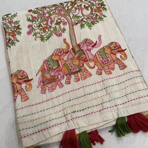 以下が含まれることがあります： 象、木、花の刺繍が施されたクリーム色の織物。象はピンク、オレンジ、緑色です。織物にはカラフルな水平ステッチと、赤、緑、黄色のタッセルが付いています。