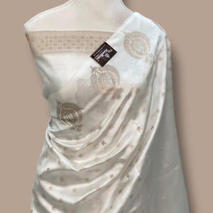 Può includere: Sari di seta color avorio con intricati ricami floreali dorati. Il sari è drappeggiato su un manichino, mostrando il suo tessuto fluido e il suo design dettagliato. Un'etichetta con testo è attaccata. Il design del sari è adatto alle occasioni speciali.