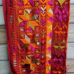 May include: A close-up of a red textile with detailed embroidery. The design incorporates geometric shapes, such as stars and arrows, in orange, yellow, pink, and silver. The embroidery is intricate and covers the entire surface of the fabric.