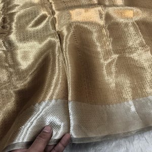 May include: Close-up of a shimmering gold fabric with a subtle woven pattern. The fabric has a metallic sheen and a contrasting silver border with a chevron design. The material is soft and appears to drape well.