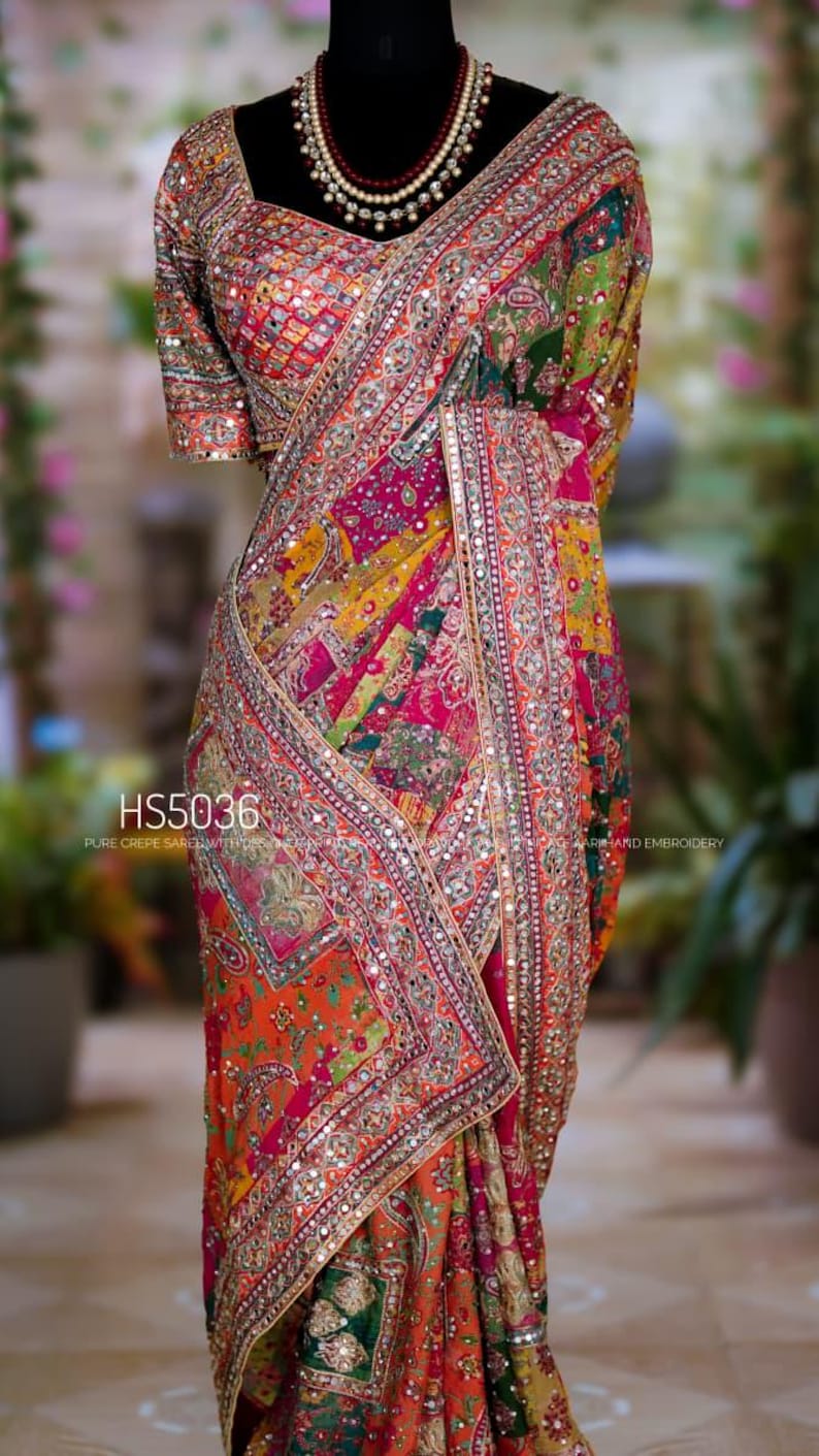 May include: A multi-colored crepe saree featuring intricate embroidery, mirror work, and a patchwork design in shades of pink, orange, green, and yellow. The garment includes a matching blouse and is labeled with the text HS5036.