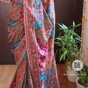 May include: A vibrant multi-coloured saree featuring a complex paisley pattern in shades of red, turquoise, orange, and green. The garment is heavily embellished with intricate silver embroidery and small reflective mirrors along the border and body.