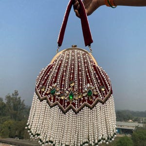 Pode incluir: Uma bolsa bordô com armação dourada e alça de veludo. A bolsa é adornada com contas de pérolas brancas e enfeites verdes e dourados. A parte inferior da bolsa tem uma franja de pérolas brancas.