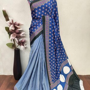 May include: A blue and white patterned saree with a striped lower section. The saree features a floral and geometric design with a dark green border. The fabric is draped on a mannequin, showcasing the intricate patterns and colors.
