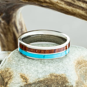 May include: A silver ring with a turquoise and wood inlay. The ring is sitting on a gray rock.