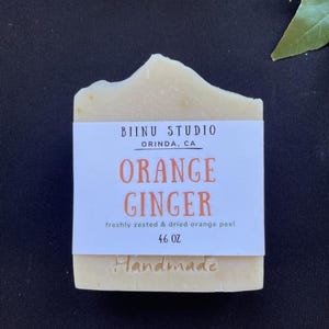 May include: A white bar of handmade soap with a label that reads "BIINU STUDIO ORINDA, CA ORANGE GINGER freshly zested & dried orange peel 4.6 OZ Handmade".