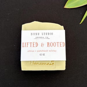 May include: A rectangular bar of light green soap wrapped in a white paper label. The label reads BIINU STUDIO ORINDA, CA LIFTED & ROOTED citrus + patchouli w/clay 4.5 OZ. The word Handmade is embossed in the soap below the paper label.