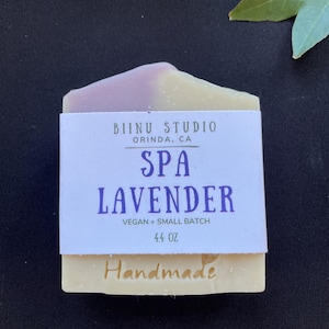 May include: A lavender-scented soap bar with a purple and white swirl. The bar is labeled "Spa Lavender" and "Handmade" with the company name "Biinu Studio, Orinda, CA". The soap is vegan and small batch, and weighs 4.4 ounces.