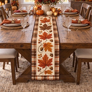 May include: A rectangular table runner featuring a central cream-colored panel with a printed pattern of orange, red, and brown maple leaves and acorns, bordered by a brown and white plaid fabric, displayed on a wooden dining table.