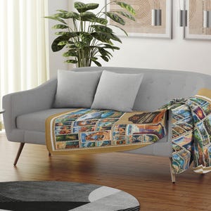 May include: A gray couch with two pillows and a throw blanket with a colorful pattern of national park stamps. The blanket is draped over the back of the couch and hangs down the side.