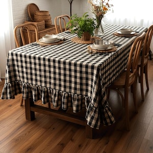 May include: A dining room with a black and white checked tablecloth on a wooden table. The tablecloth has a frilled edge. The table is set with plates and placemats. Wicker chairs surround the table, and a woven pendant light hangs above.