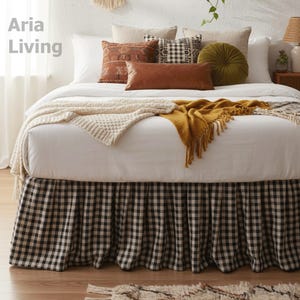 Może przedstawiać: Łóżko z białą pościelą, kremowym dzianinowym kocem i musztardową frędzlą. Ozdobne poduszki w różnych fakturach i kolorach, w tym brązowa skóra i oliwkowa zieleń, zdobią łóżko. Spódnica łóżka ma czarno-biały wzór w kratę. Widoczny jest tekst "Aria Living".
