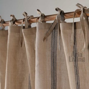 Puede incluir: Cortinas de lino beige con rayas verticales azul oscuro y lazos. Las cortinas cuelgan de una barra de madera. El texto "Atia Living" es visible en el lado derecho de las cortinas.
