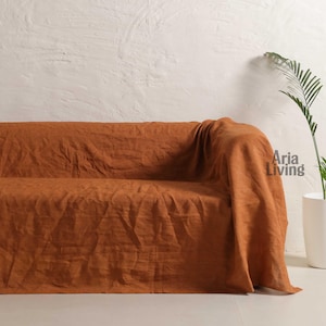 Op de afbeelding: Een roestkleurige linnen bankhoes over een bank gedrapeerd. De hoes heeft een textuur en is gemaakt van een zachte, vloeiende stof. De bank staat tegen een witte muur, met een plant in een pot op de achtergrond. De tekst "Aria Living" is zichtbaar.