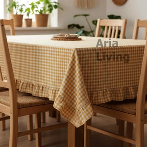 May include: A yellow and white checkered tablecloth with a ruffled edge covers a wooden table. The table is surrounded by wooden chairs with woven seats. The text "Aria Living" is visible on the tablecloth.