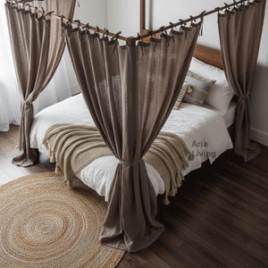 Può includere: Una scena di camera da letto con un letto con struttura in legno e un baldacchino di tende in lino marrone. Il letto ha lenzuola bianche, una coperta beige e cuscini decorativi. Un tappeto rotondo in juta è sul pavimento in legno.