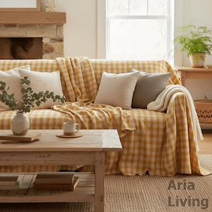 Può includere: Un soggiorno con un divano coperto da una coperta a scacchi gialli e bianchi. Diversi cuscini dai toni neutri e una coperta color crema adornano il divano. Un tavolino in legno espone libri, una tazza e un sottobicchiere. È presente anche un vaso con vegetazione.