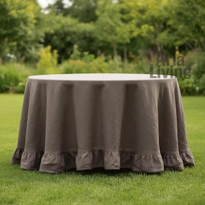 Puede incluir: Un mantel redondo marrón con borde con volantes, colocado sobre una mesa blanca. El mantel está hecho de una tela texturizada y está al aire libre sobre un césped. El fondo presenta árboles y follaje verdes.