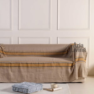 May include: A beige sofa draped with a striped throw in yellow and dark blue. A striped cushion and a cup of coffee with a book are on the floor. The wall behind the sofa is white with panel detailing.