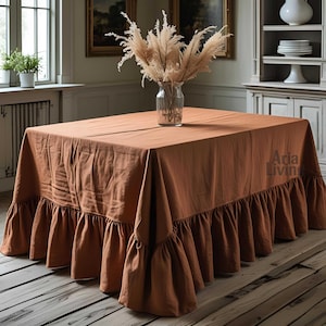 May include: A square, rust-colored tablecloth with a ruffled edge. The tablecloth is draped over a wooden table, and a clear glass vase with dried flowers sits in the center. The Aria Living logo is visible on the tablecloth.