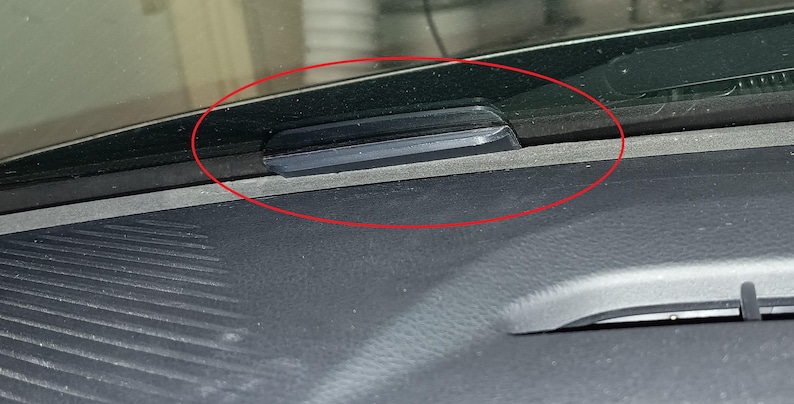 Op de afbeelding: Een zwart, rechthoekig object is gemonteerd op het dashboard van een voertuig, in de buurt van de voorruit. Het object lijkt gemaakt te zijn van een glad, donker materiaal en is horizontaal geplaatst. Het dashboard is donkergrijs.