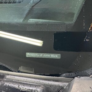 Op de afbeelding: Een close-up van een autoruit met de tekst "Property of John Wick" op een kleine rechthoekige sticker. De voorruit heeft een rasterpatroon aan de bovenkant.