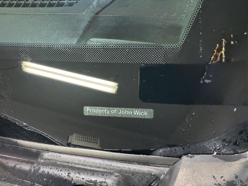 Op de afbeelding: Een close-up van een autoruit met de tekst "Property of John Wick" op een kleine rechthoekige sticker. De voorruit heeft een rasterpatroon aan de bovenkant.