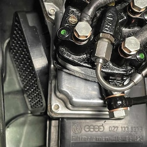 Op de afbeelding: Close-up van een zwart auto-onderdeel met metalen fittingen en een zilveren buis. Het onderdeel heeft zeskantbouten en de tekst "G895 027 133 837F". Gedetailleerde weergave van het motoronderdeel.