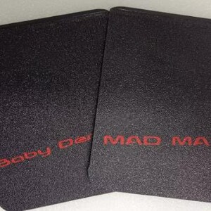Op de afbeelding: Twee zwarte rechthoekige items met een getextureerd oppervlak. De tekst "Baby Der" is rood gedrukt op het linker item en "MAD MAX" is rood gedrukt op het rechter item.