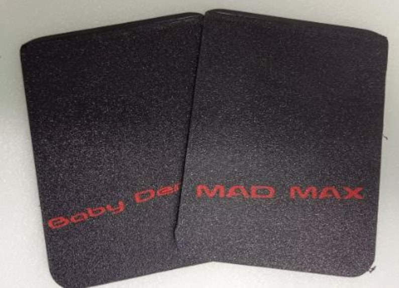 Op de afbeelding: Twee zwarte rechthoekige items met een getextureerd oppervlak. De tekst "Baby Der" is rood gedrukt op het linker item en "MAD MAX" is rood gedrukt op het rechter item.