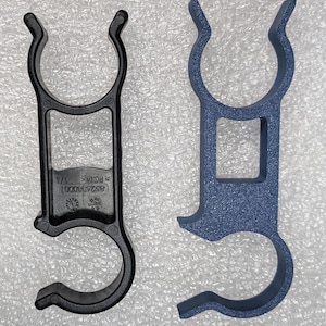 May include: Two plastic clips, one black and one blue, each featuring a dual-ring design with a central square opening. The black clip displays molded text reading 8526380001, POM, and 1/1, along with a small circular indicator symbol on its surface.
