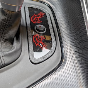 Op de afbeelding: Detail van het interieur van een auto met een zwart en zilveren console. Een zwart paneel met een rood logo en een knop is zichtbaar. De versnellingspook is van zwart leer. De console heeft een gestructureerd oppervlak.