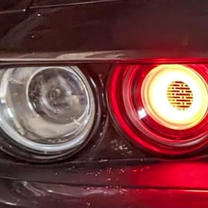 Op de afbeelding: Close-up van de koplamp en het achterlicht van een auto. De koplamp is helder, terwijl het achterlicht rood oplicht met een heldergeel centrum. De carrosserie van de auto is donker en de lichten zijn ingeschakeld.